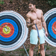 Shirtless man standing in front of 2 large archery targets holding a bow and wearing white mesh shorts.