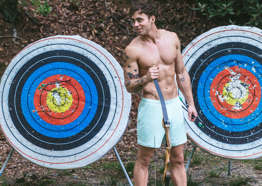 Shirtless man standing in front of 2 large archery targets holding a bow and wearing white mesh shorts.