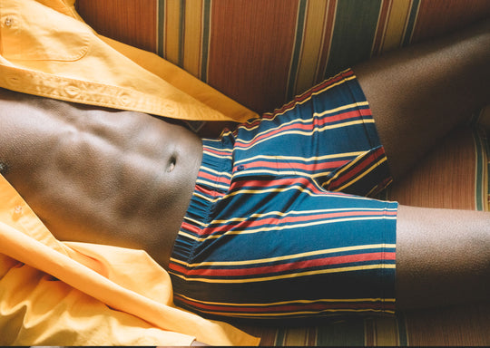 Close up shot of man sitting on couch wearing open yellow dress shirt and green/blue/red stripe slim fit boxers.