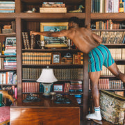 Man standing in library grabbing a book wearing green/blue/yellow stripe slim fit boxers.