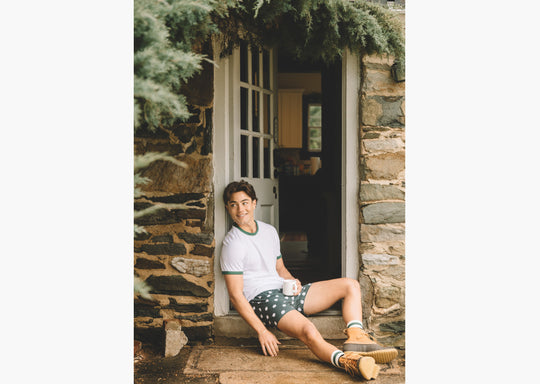 Man sitting on porch holding coffee mug and wearing white shirt, boots and green slim fit boxers with white dots.