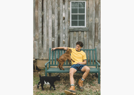 Man sitting on bench with small goat. Man is wearing yellow shirt, boots and blue slim fit boxers with white dots.