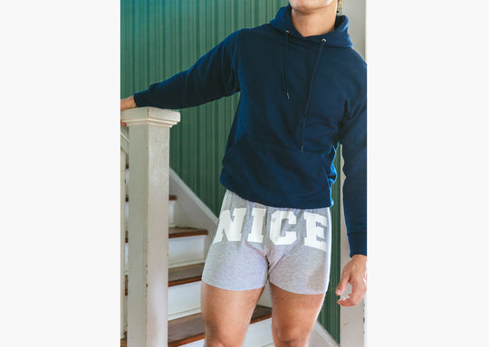 Man standing inside of house on stairs wearing navy blue sweatshirt and grey slim fit boxers with words "NICE" on the front in white bold font.
