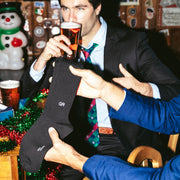 Black socks with a white GR monogram held in front of a man drinking beer at a holiday-decorated bar.