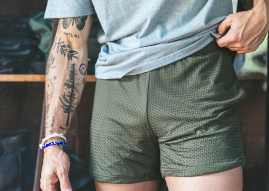 Man with tattoos wearing grey shirt and olive mesh shorts.