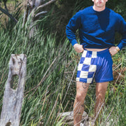 Man standing in bushes by beach wearing blue sweater and checkered/solid navy blue lounge shorts.