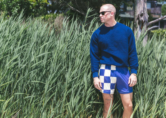 Man standing next to bushes wearing half checkered half solid blue and white mesh lounge shorts.