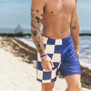 Shirtless man walking down the beach wearing half checkered half solid blue and white mesh lounge shorts.