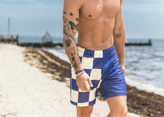 Shirtless man walking down the beach wearing half checkered half solid blue and white mesh lounge shorts.