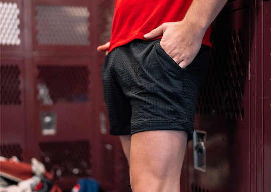 Close up of man in locker room wearing black lounge shorts and red shirt with his hand in pocket.