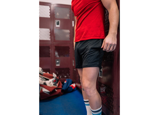 Man in locker room wearing red shirt, black lounge shorts, and black/blue stripe crew socks.