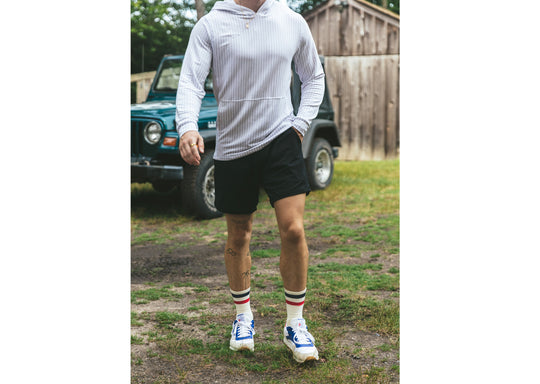 Man standing in front of blue jeep wearing light linen stripe hoodie, black lounge shorts, and blue/red stripe varsity socks.