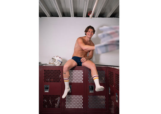 Shirtless man sitting on locker wearing blue boxer brief and orange/red stripe crew socks.