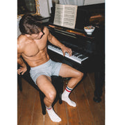 Shirtless man sitting at a piano wearing grey slim fit boxers and blue/red stripe tech varsity socks.