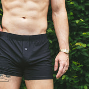 Shirtless man standing in front of bush wearing black slim fit boxers with his hand on hip.