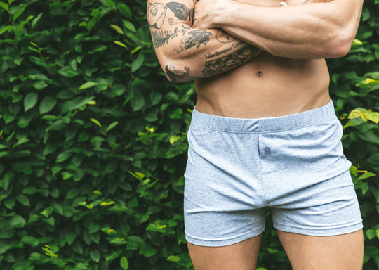 Shirtless man standing arms crossed in front of bush wearing gray slim fit boxer.