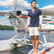 A man stands barefoot on the front float of a white seaplane on water under a blue sky. He is wearing a navy blue henley shirt with yellow and white trim, white long-sleeve undershirt, and white shorts.