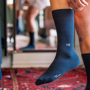 Close up of man lifting his leg to put on navy blue ribbed dress sock with VB initials monogrammed on one.
