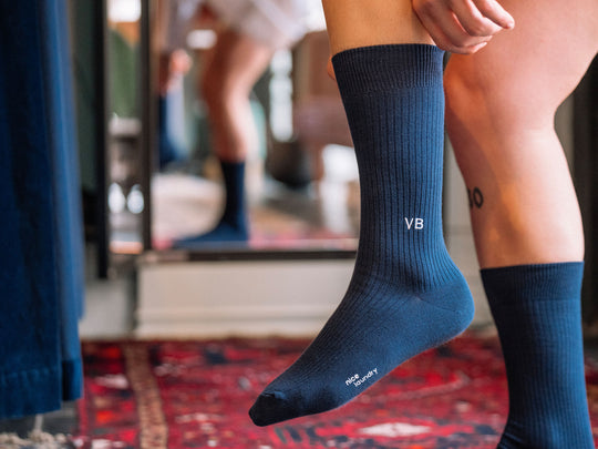 Close up of man lifting his leg to put on navy blue ribbed dress sock with VB initials monogrammed on one.