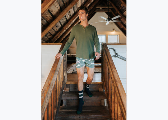 Man walking down stairs wearing long sleeve green t-shirt and green slim fit boxers with clouds, horses and trees printed on.