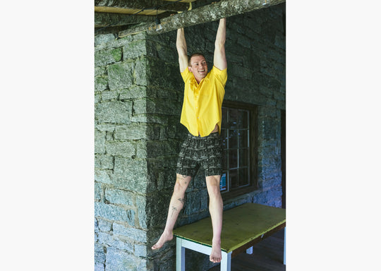 Man hanging in the air holding on to a wooden beam wearing a yellow polo and black "schooner" mesh lounge shorts.