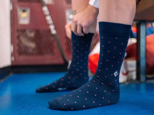 Man with both foot on the ground getting dressed wearing blue dot dress socks with initials "JBA" on one.