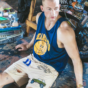 Man sitting leaning against a motorcycle in a garage wearing senior cord lounge shorts and blue tank.