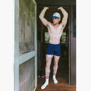 Shirtless man standing in doorway of cabin wearing navy blue slim fit boxers and blue/white cap.