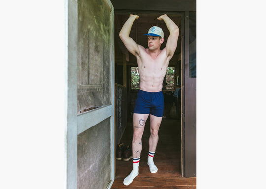 Shirtless man standing in doorway of cabin wearing navy blue slim fit boxers and blue/white cap.