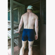 Back shot of a shirtless man standing in doorway of cabin wearing navy blue slim fit boxers and white/blue cap.