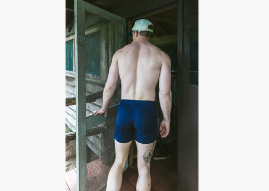 Back shot of a shirtless man standing in doorway of cabin wearing navy blue slim fit boxers and white/blue cap.
