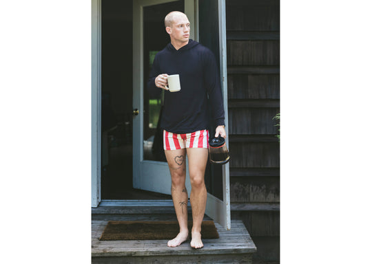 Man wearing black hoodie and red/cream stripe slim fit boxing standing outside front door holding coffee mug.
