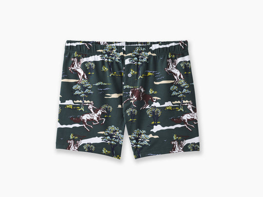 Pair of hunter green slim fit boxers with trees and men riding horses printed on sitting against light grey background.