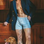 A shirtless man wearing a navy blazer and light blue striped boxer briefs stands in a wood-paneled room with a golden retriever dog at his side.