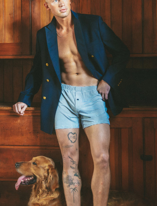 A shirtless man wearing a navy blazer and light blue striped boxer briefs stands in a wood-paneled room with a golden retriever dog at his side.