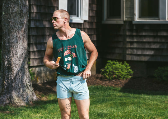 Man wearing sleeveless black shirt and green striped slim fit boxers standing outside in front of a house.