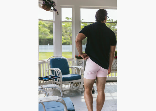 Back shot of man standing inside wearing black polo shirt and pink striped slim fit boxers.