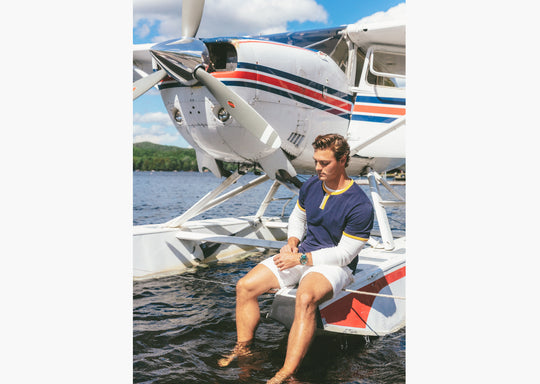 Man sitting on dock in front of plane wearing Navy vintage ringer and cream lounge shorts