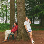 Two men in a forest setting, one sitting on a teal chair wearing a red hoodie and white pants, and the other leaning against a large tree wearing a white t-shirt, blue and white striped shorts, and white socks.