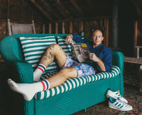 A man reclining on a teal woven outdoor sofa, holding a magazine. He is wearing a blue long-sleeve shirt, plaid shorts, white socks with red and blue stripes, and white sneakers with blue and yellow accents.