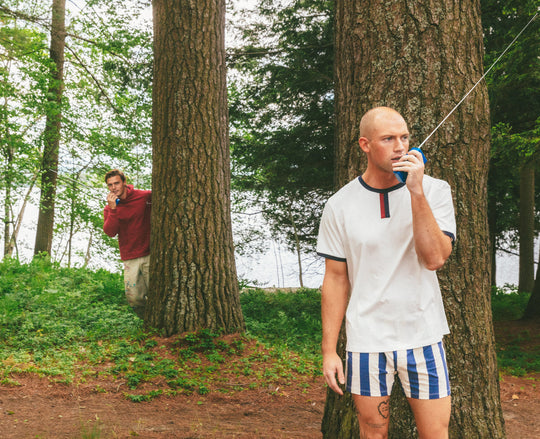 A man in a white t-shirt with blue and red trim and blue-and-white striped shorts stands in a wooded outdoor setting, holding a small blue and red radio to his mouth. Another man in a red shirt is partially hidden behind a tree in the background.