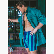 Man with green jacket and green/navy striped lounge shorts standing in front of a window and holding a fishing pole