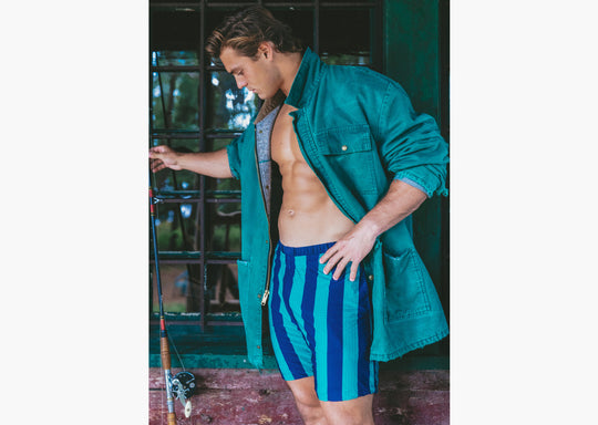 Man with green jacket and green/navy striped lounge shorts standing in front of a window and holding a fishing pole