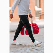 Man crossing cobblestone street wearing black lounge shorts with red bag.